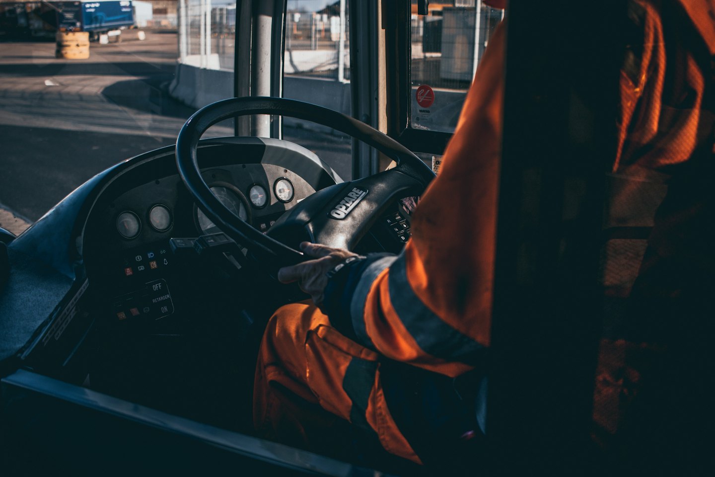 Driver in orange uniform grips the steering wheel of a large vehicle.
